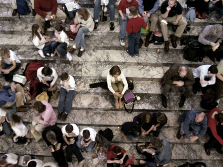 A woman with ADHD, feeling isolated from a group of people, sits on a staircase and clutches her knees to her chest.