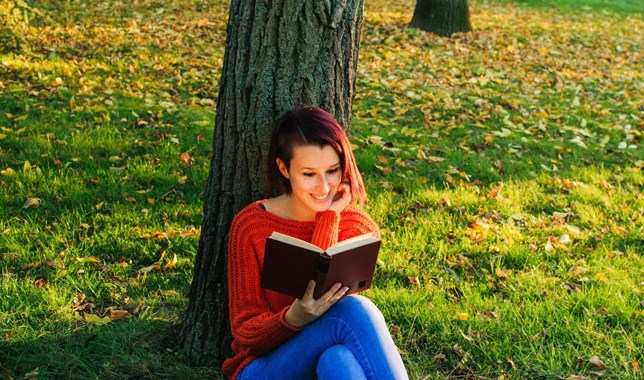Young woman sitting under a tree reading stories to help her escape cancer trauma